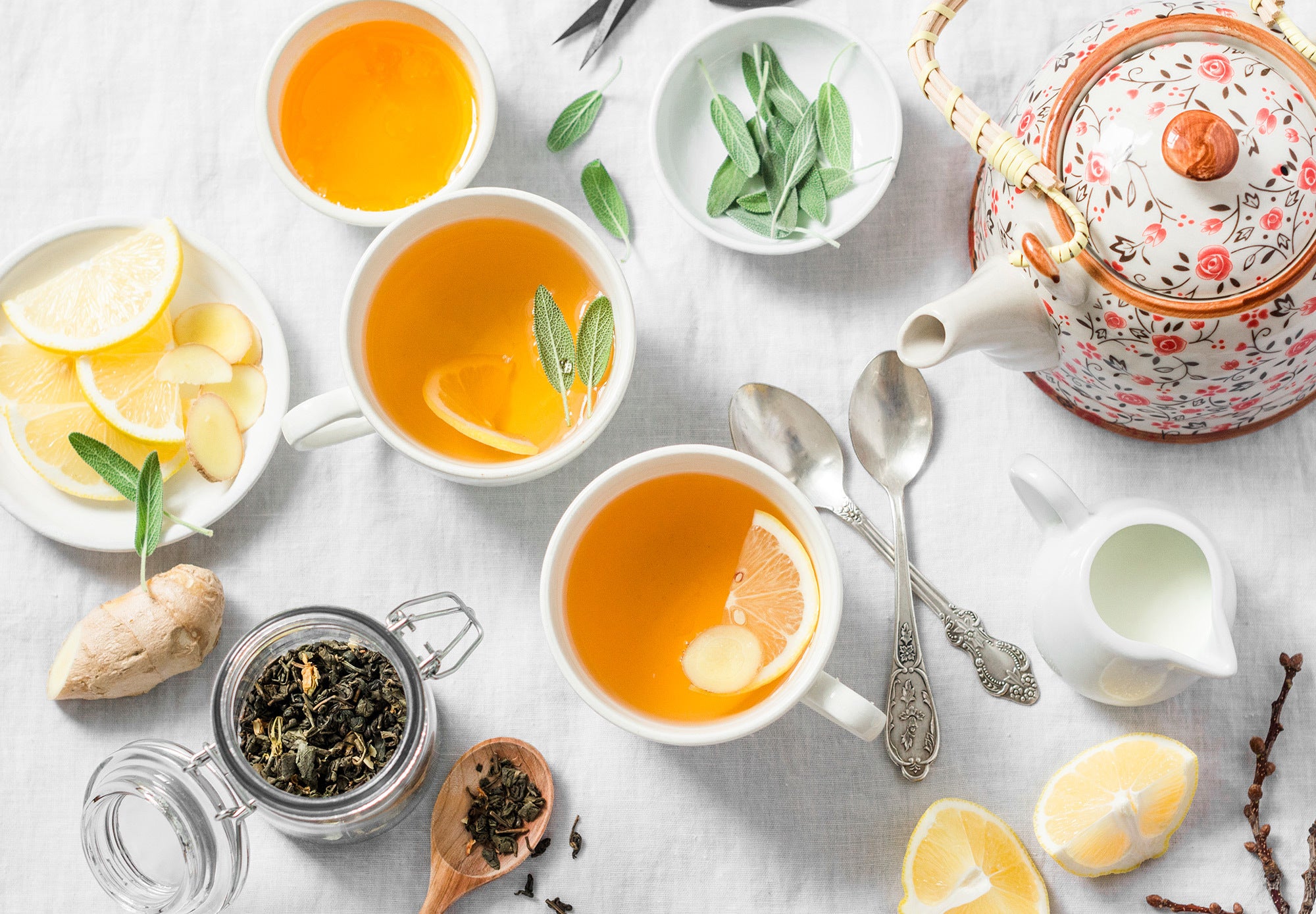 Cups of tea n a white tablecloth with fresh sage, lemon slices, ginger, cream and a teapot