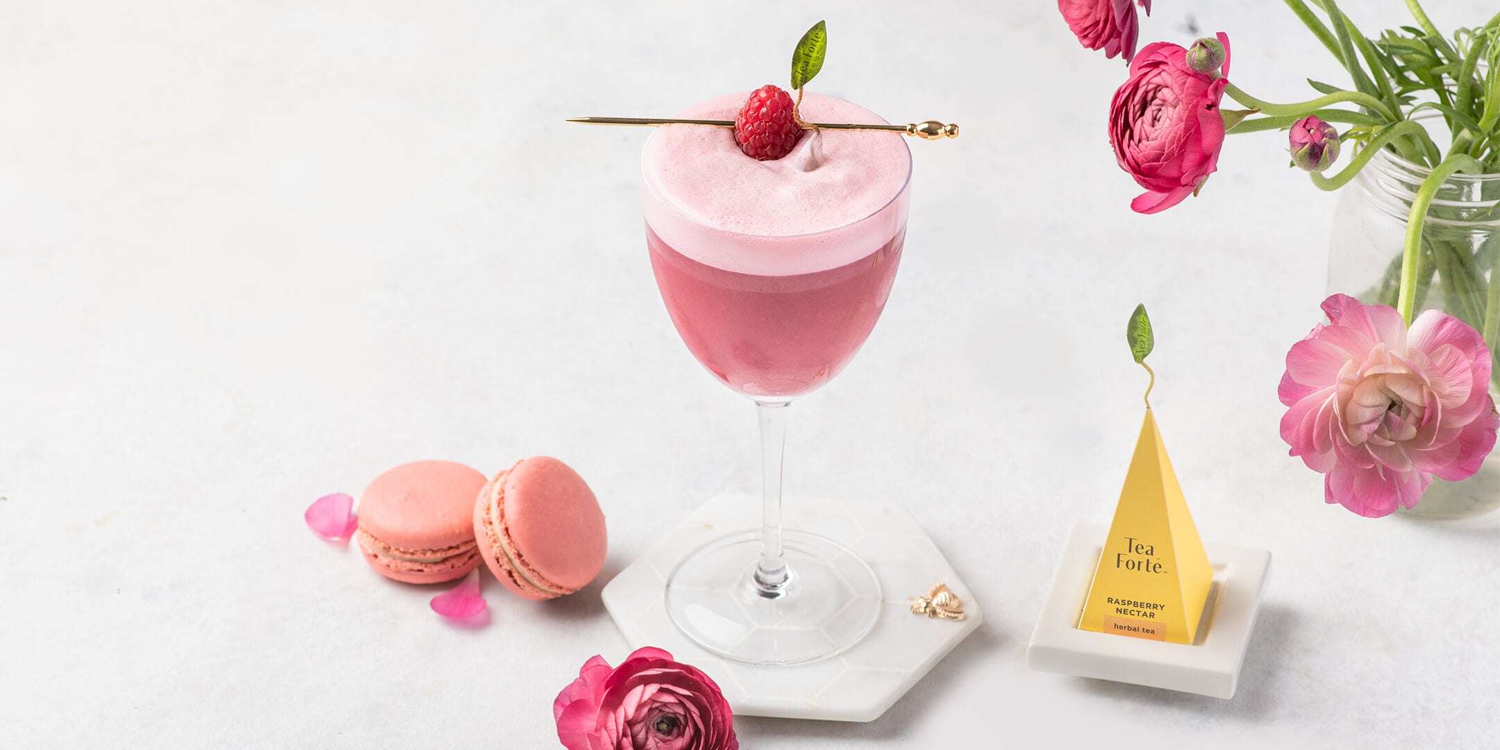 Raspberry Nectar Clover Club cocktail on a white marble tabletop with a tea infuser, pink flowers and macarons