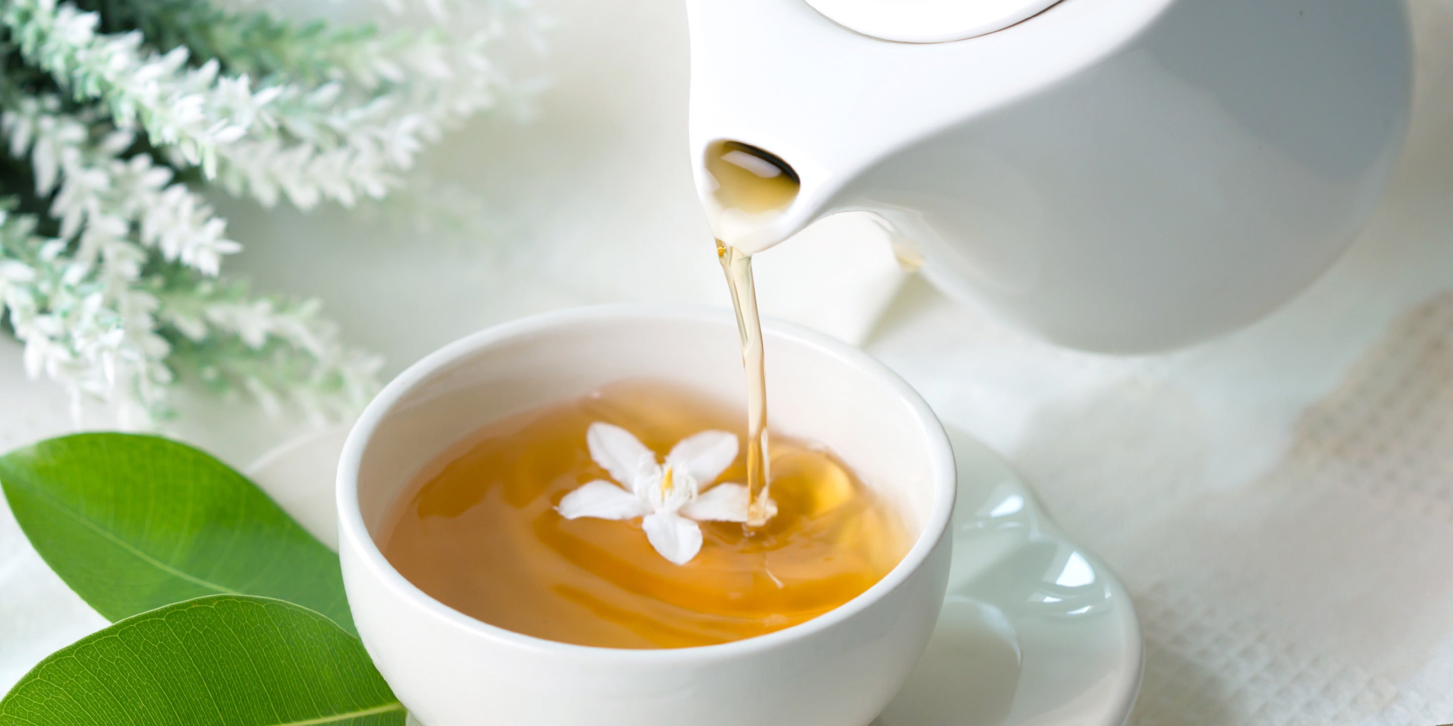 Pouring tea into a white cup from a white teapot, with a jasmine flower floating in the cup